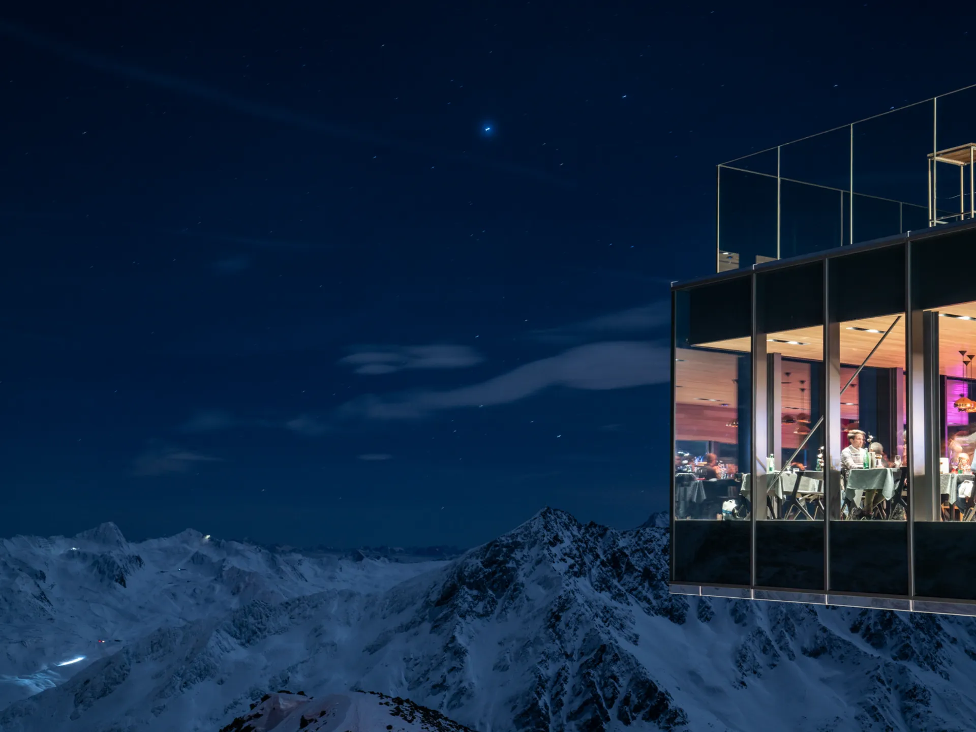 Beleuchtetes Bergrestaurant mit Gästen über schneebedeckten Gipfeln bei Nacht