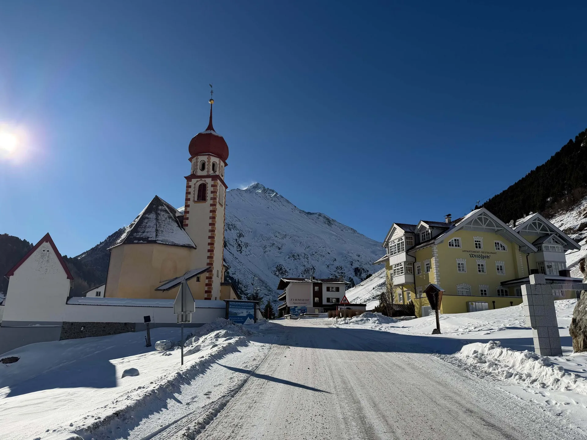 Verschneites Alpendorf mit Kirche und gelbem Hotel vor schneebedeckten Bergen