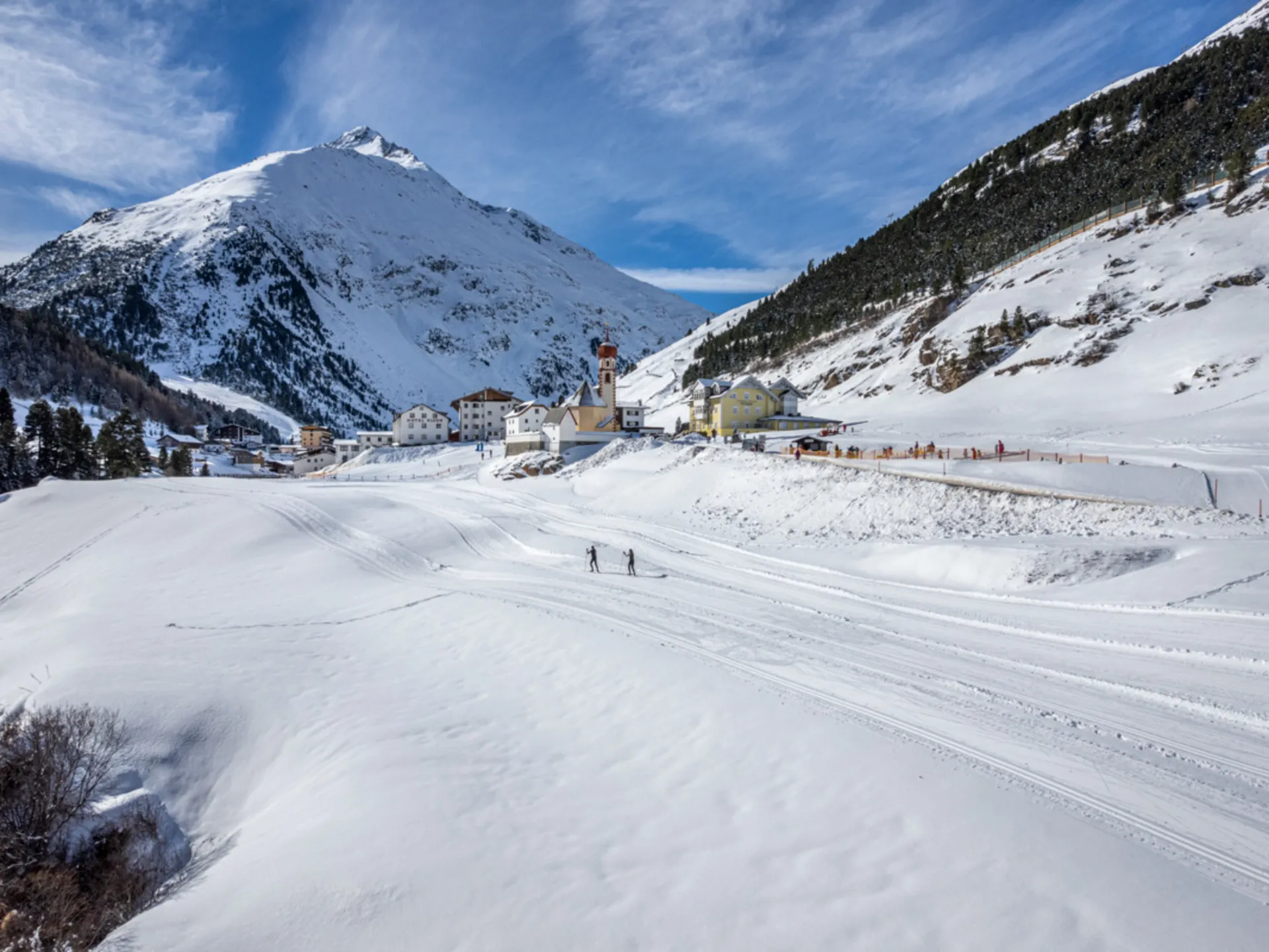 Zwei Langläufer auf einer verschneiten Loipe vor einem Bergdorf in den Alpen