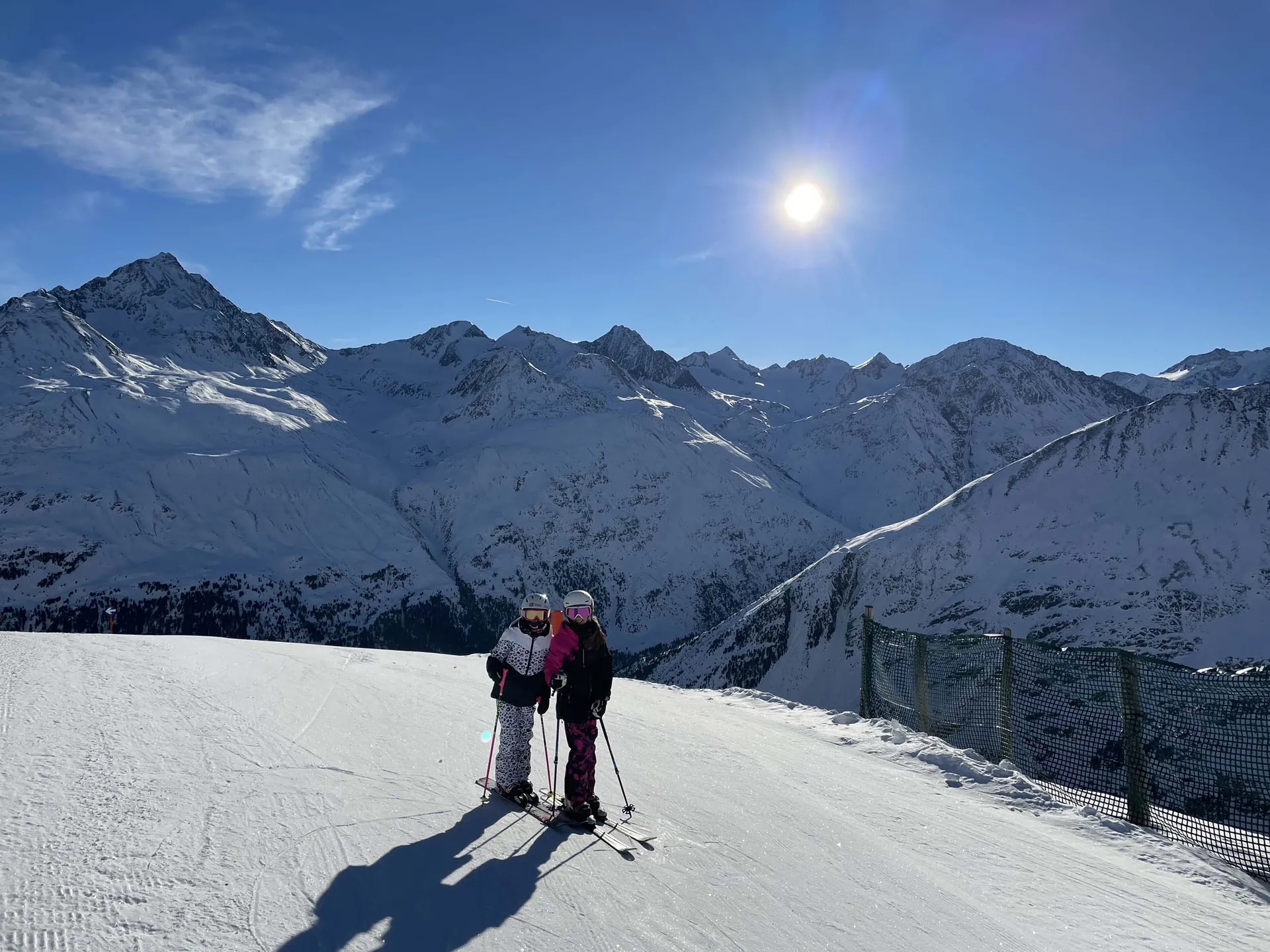 Zwei Skifahrerinnen auf einer sonnigen Piste vor einer verschneiten Berglandschaft
