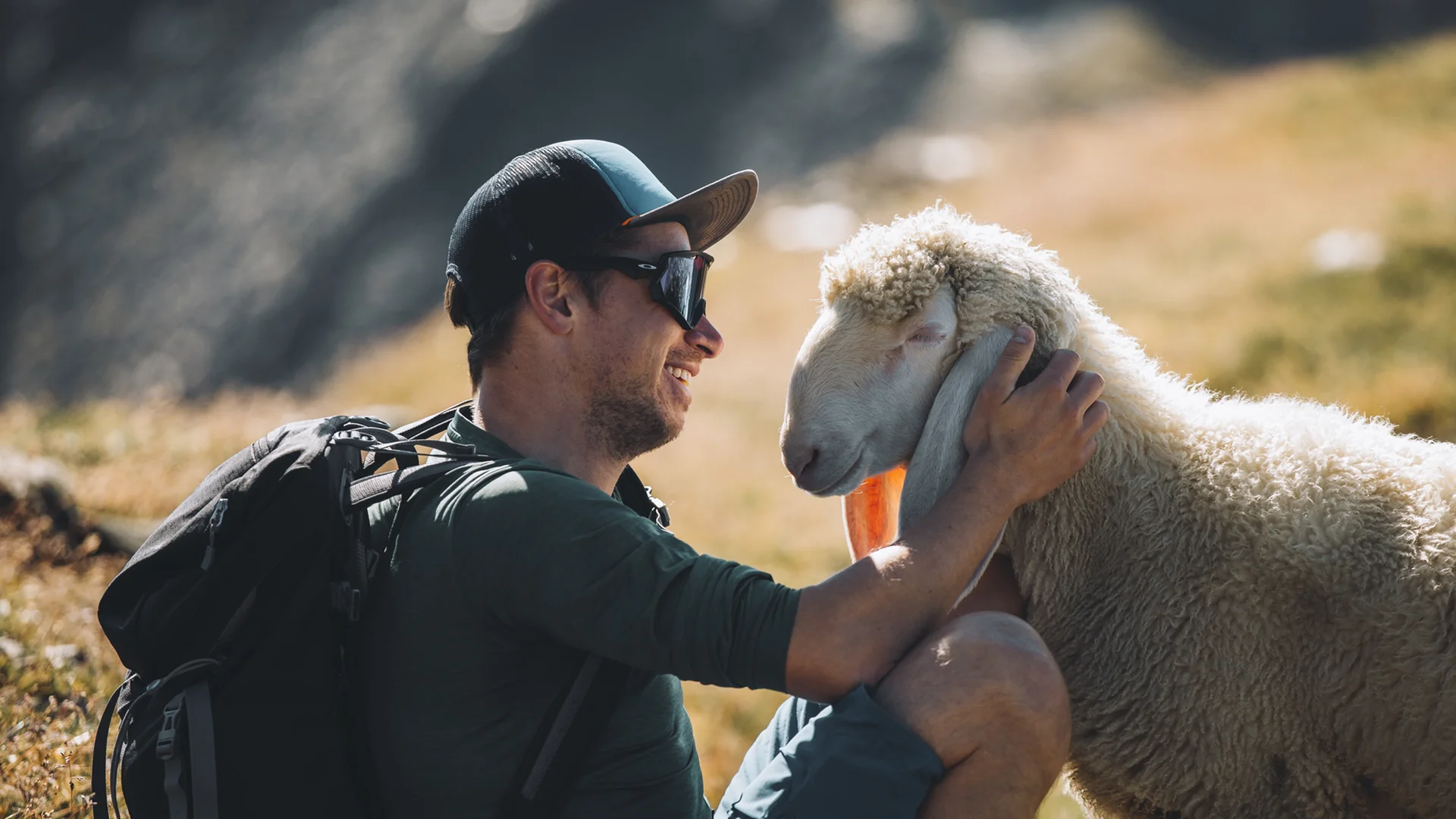 Mann mit Rucksack streichelt ein Schaf auf einer Bergwiese