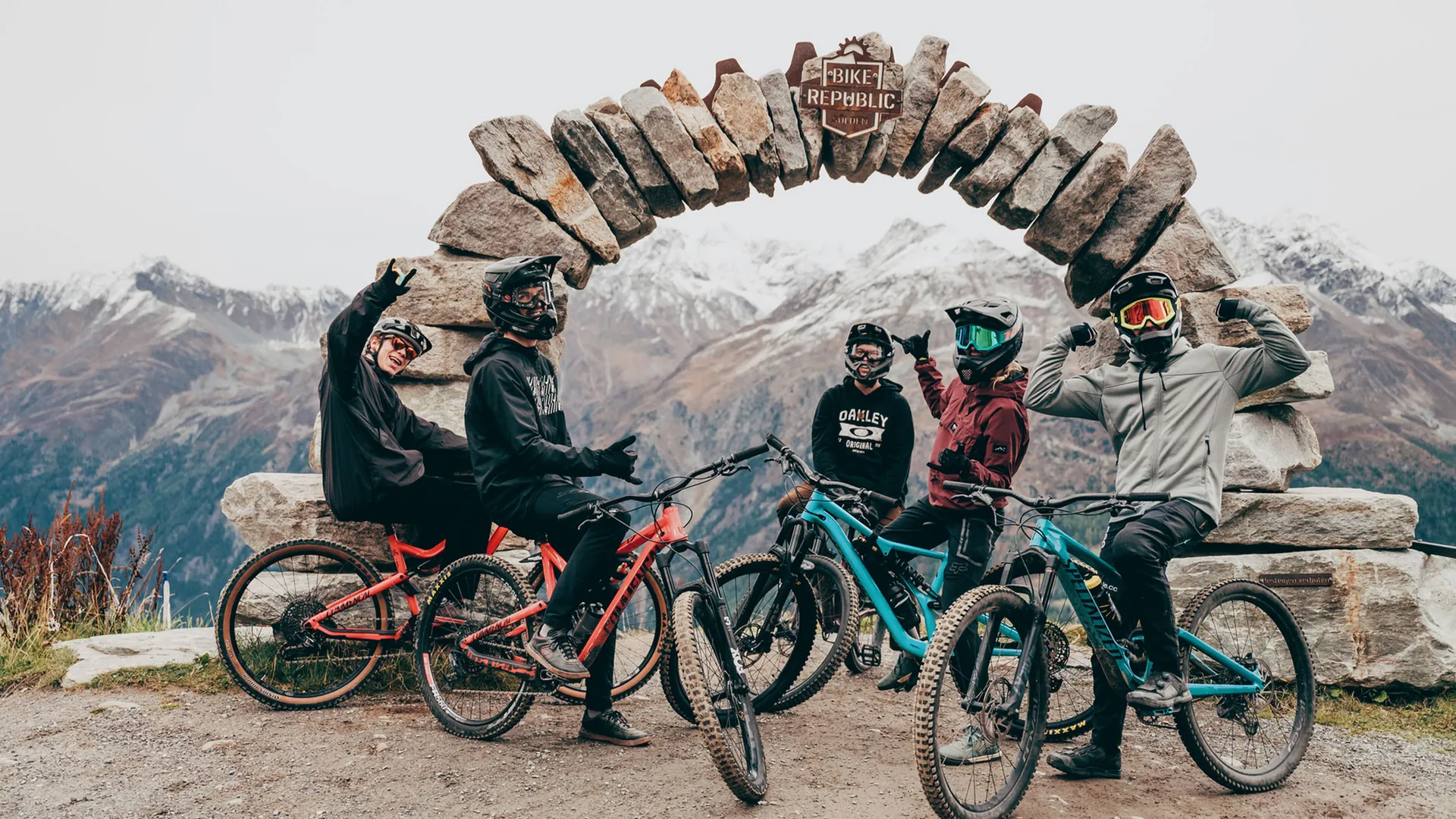 Five mountain bikers beneath a stone arch with snow-capped mountains