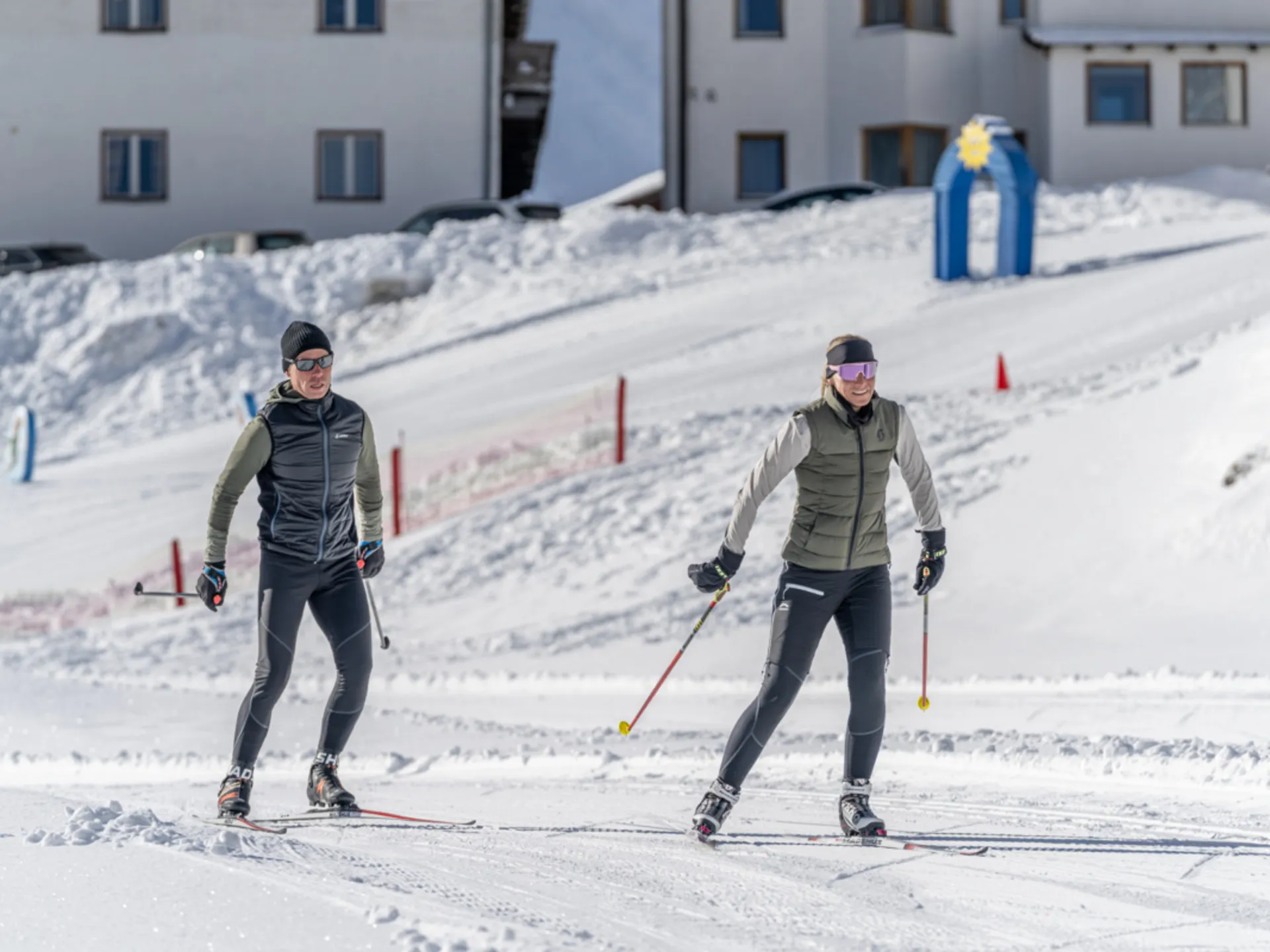 Zwei Langläufer auf einer verschneiten Loipe vor Gebäuden