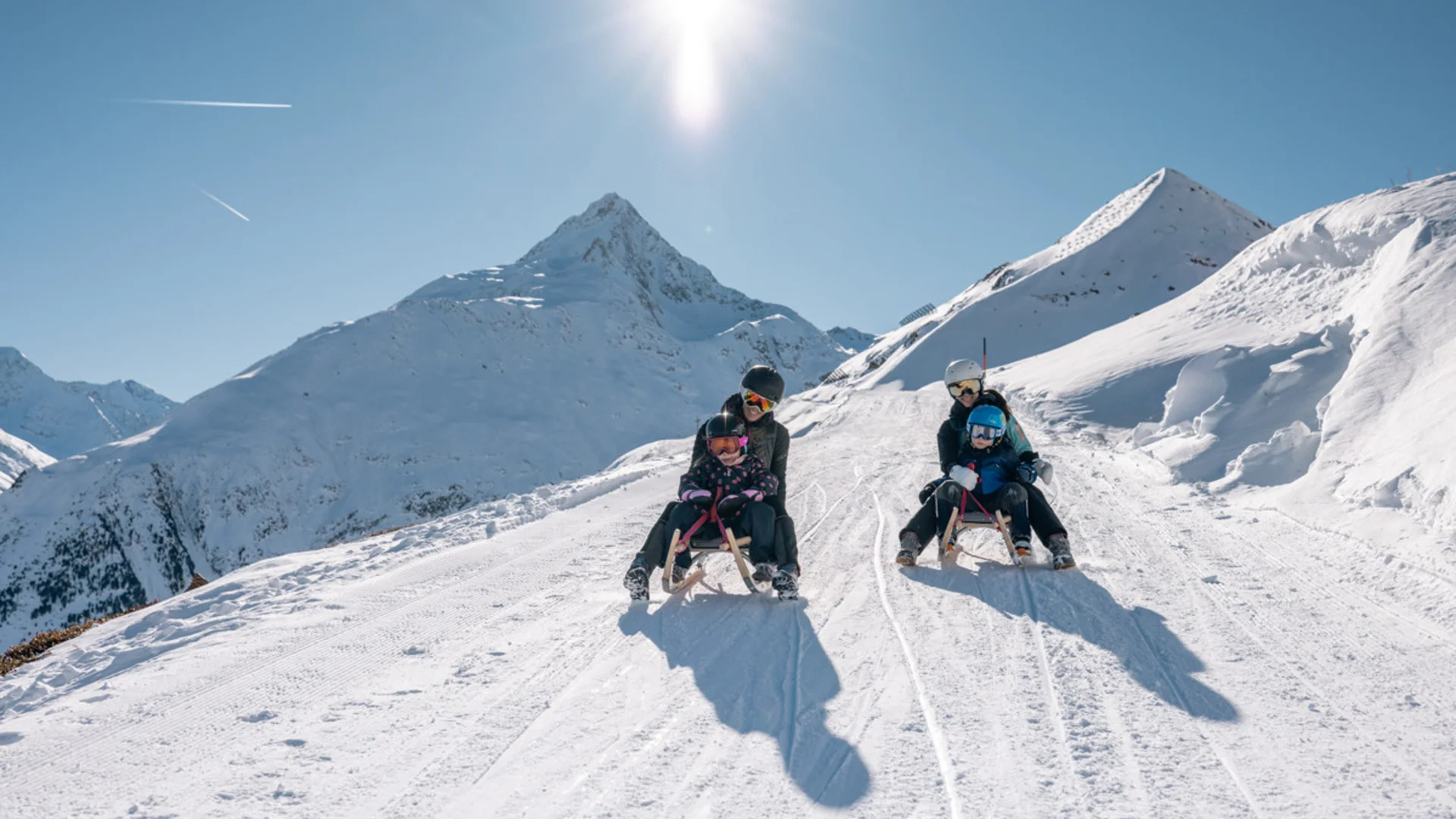 Zwei Erwachsene mit Kindern rodeln auf verschneitem Berghang bei Sonnenschein