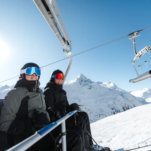 Snowy adventures on winter holidays in the Alps Zwei Skifahrer in Skikleidung auf einem Sessellift mit verschneiten Bergen im Hintergrund