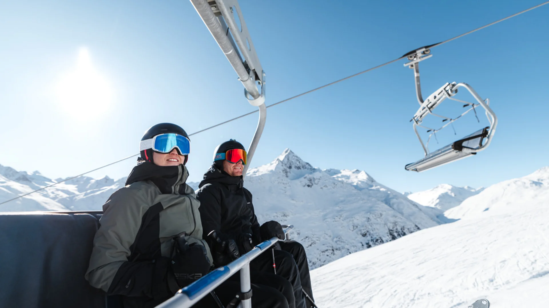 Zwei Skifahrer in Skikleidung auf einem Sessellift mit verschneiten Bergen im Hintergrund