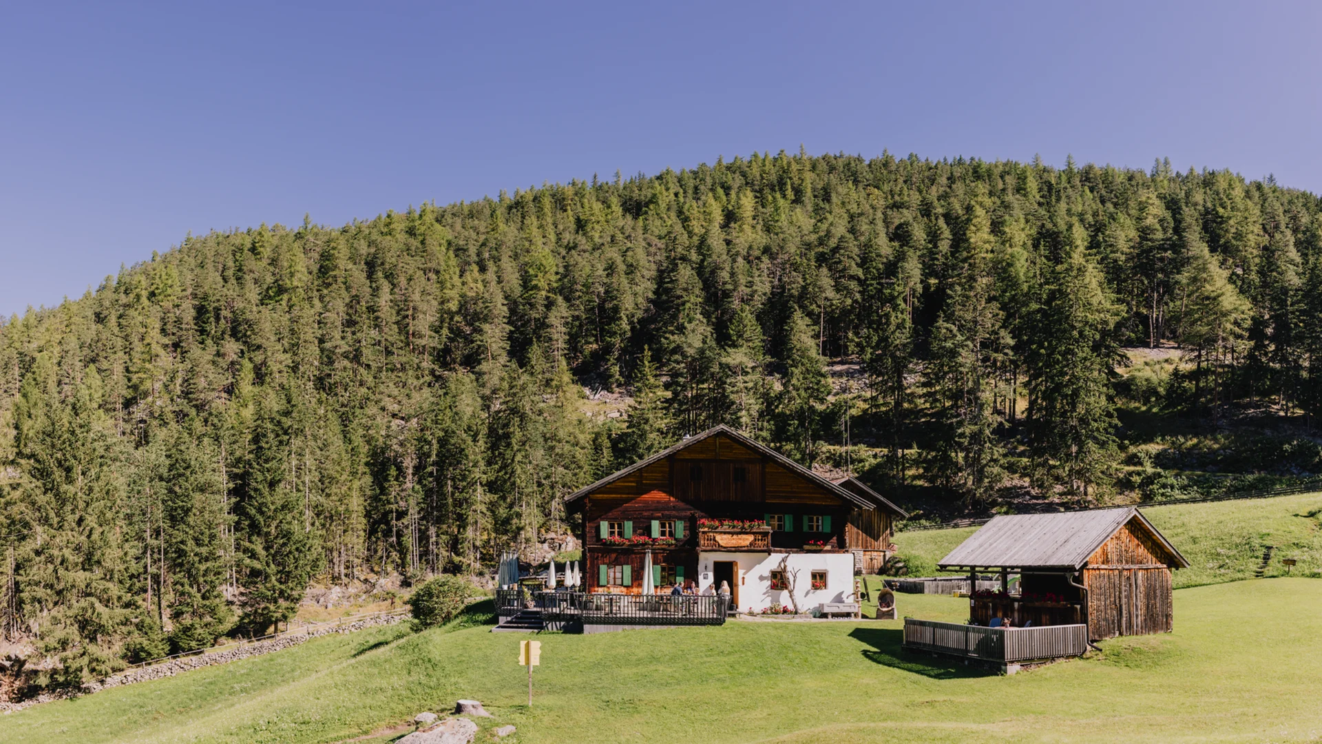 Altes Bauernhaus mit Terrasse vor bewaldetem Hügel unter klarem Himmel