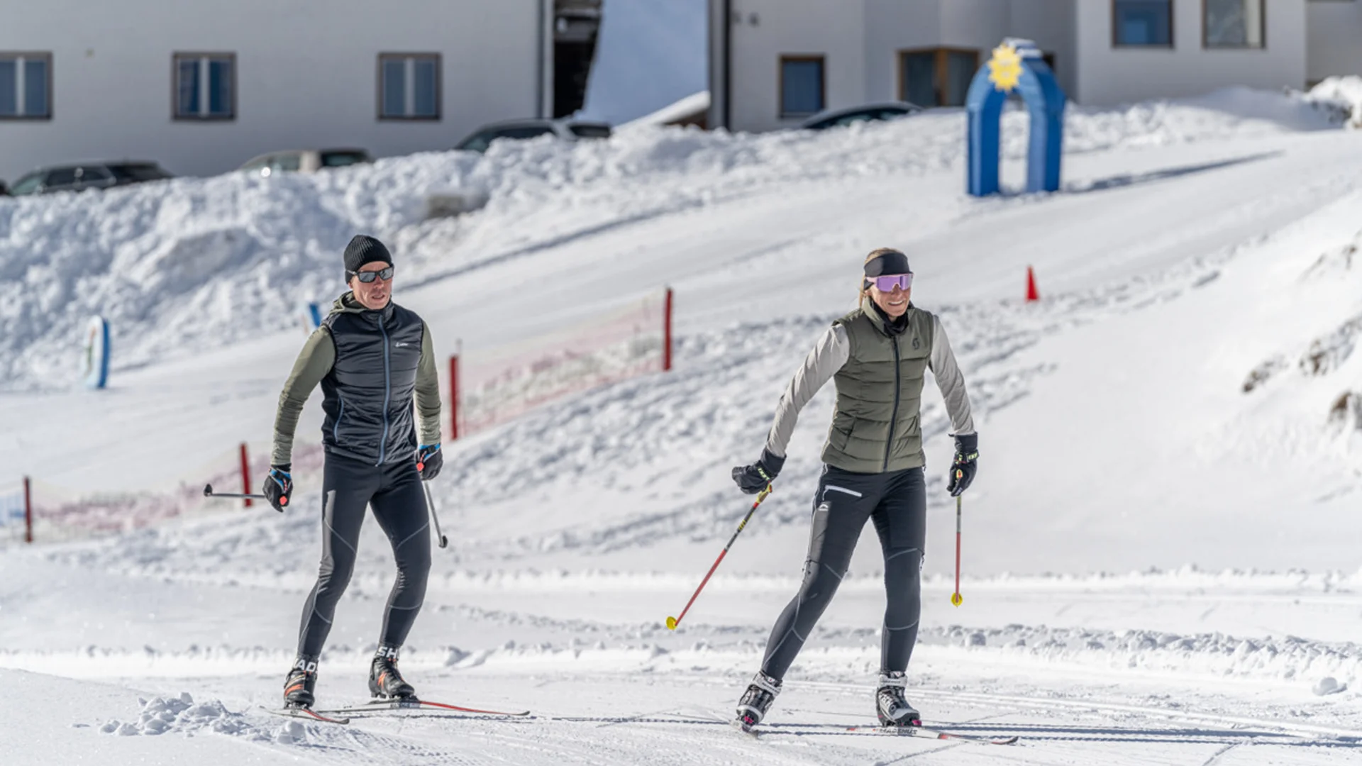 Zwei Personen beim Skilanglauf auf verschneiter Strecke vor Gebäuden