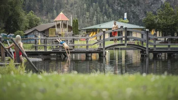 Mann springt von Holzbrücke in See, andere Personen am Ufer und auf Brücke