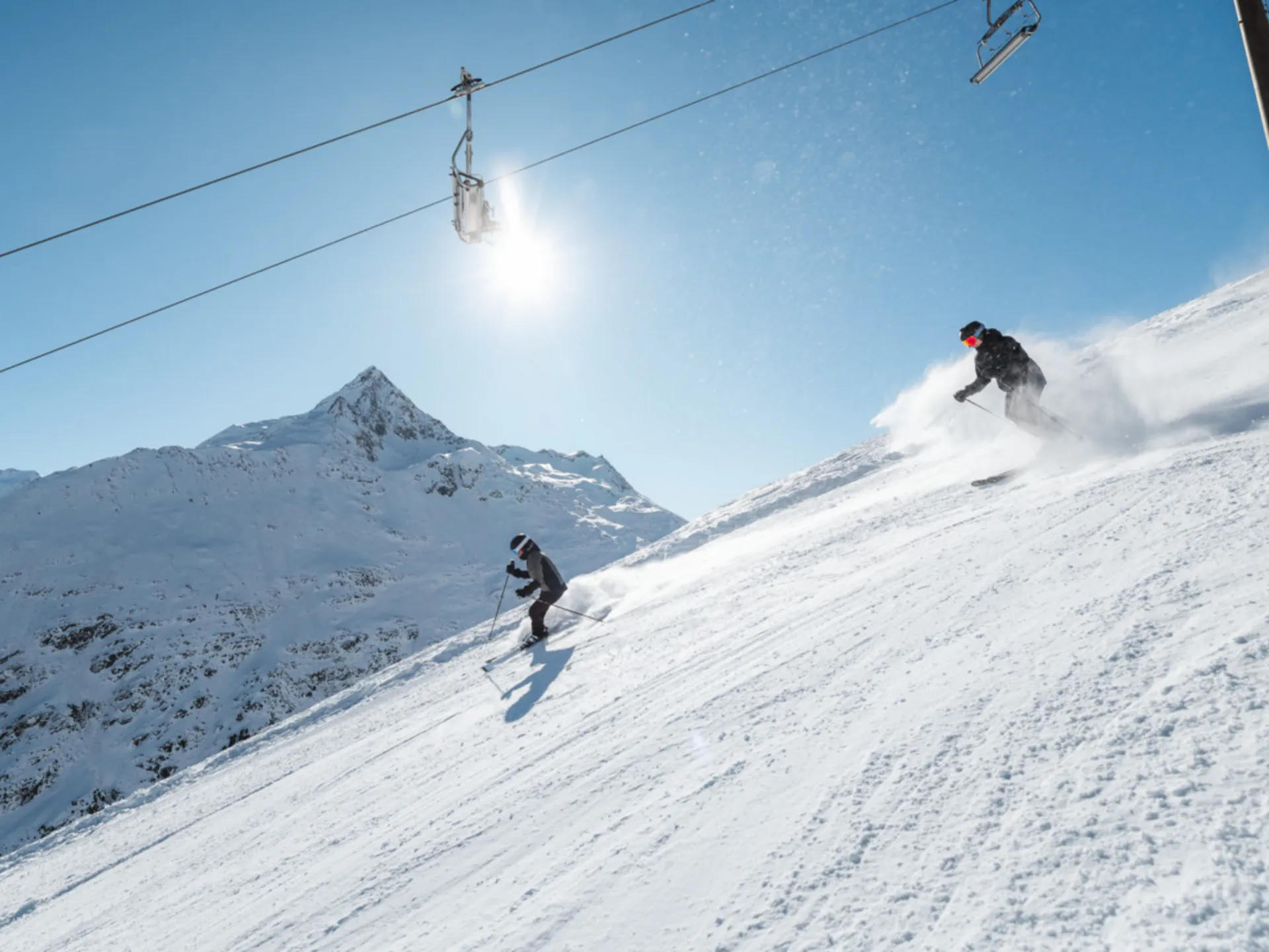Zwei Skifahrer fahren einen sonnigen Hang hinunter, über ihnen verläuft ein Skilift