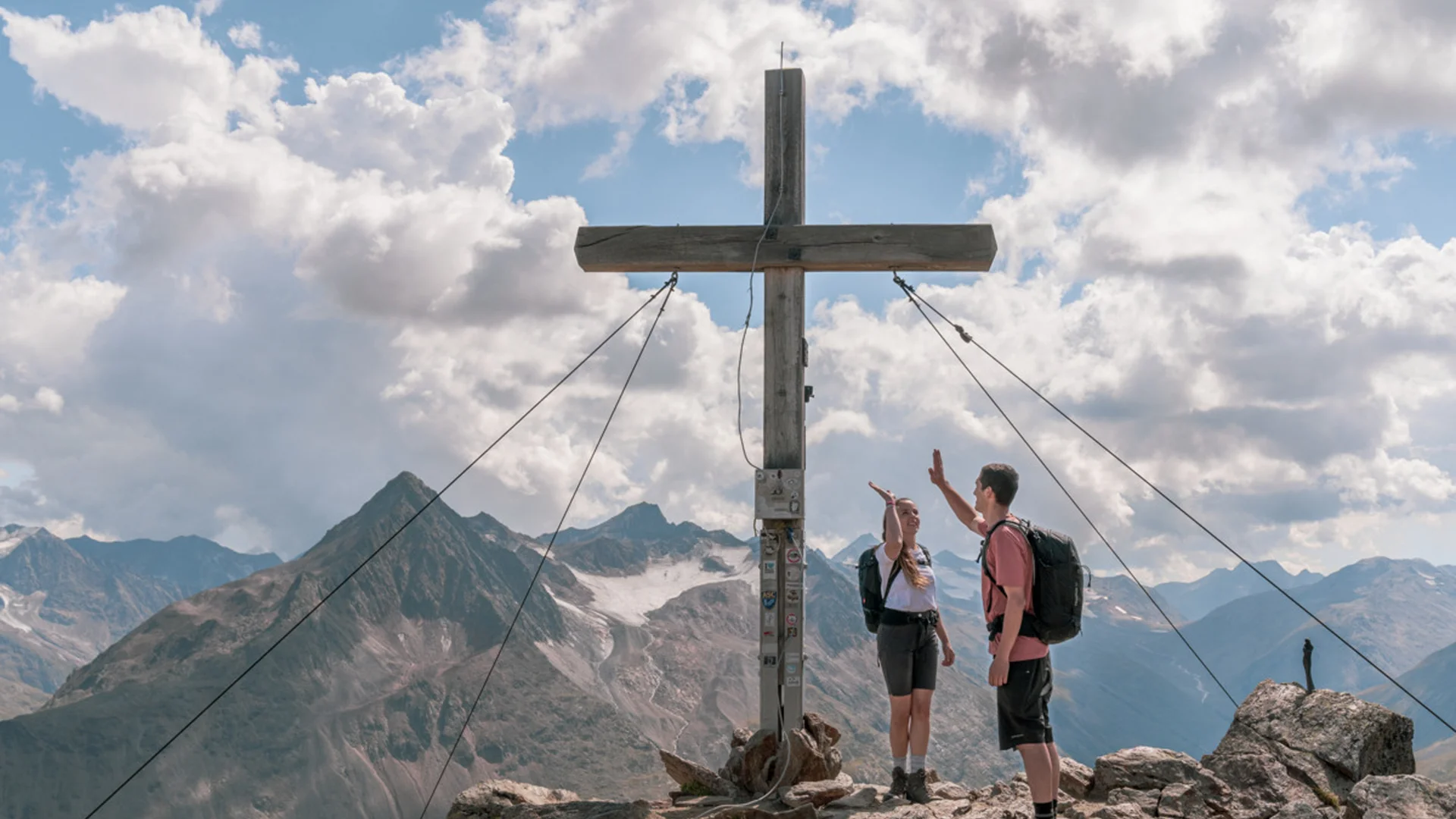 Zwei Wanderer geben sich auf Berggipfel neben großem Holzkreuz High Five