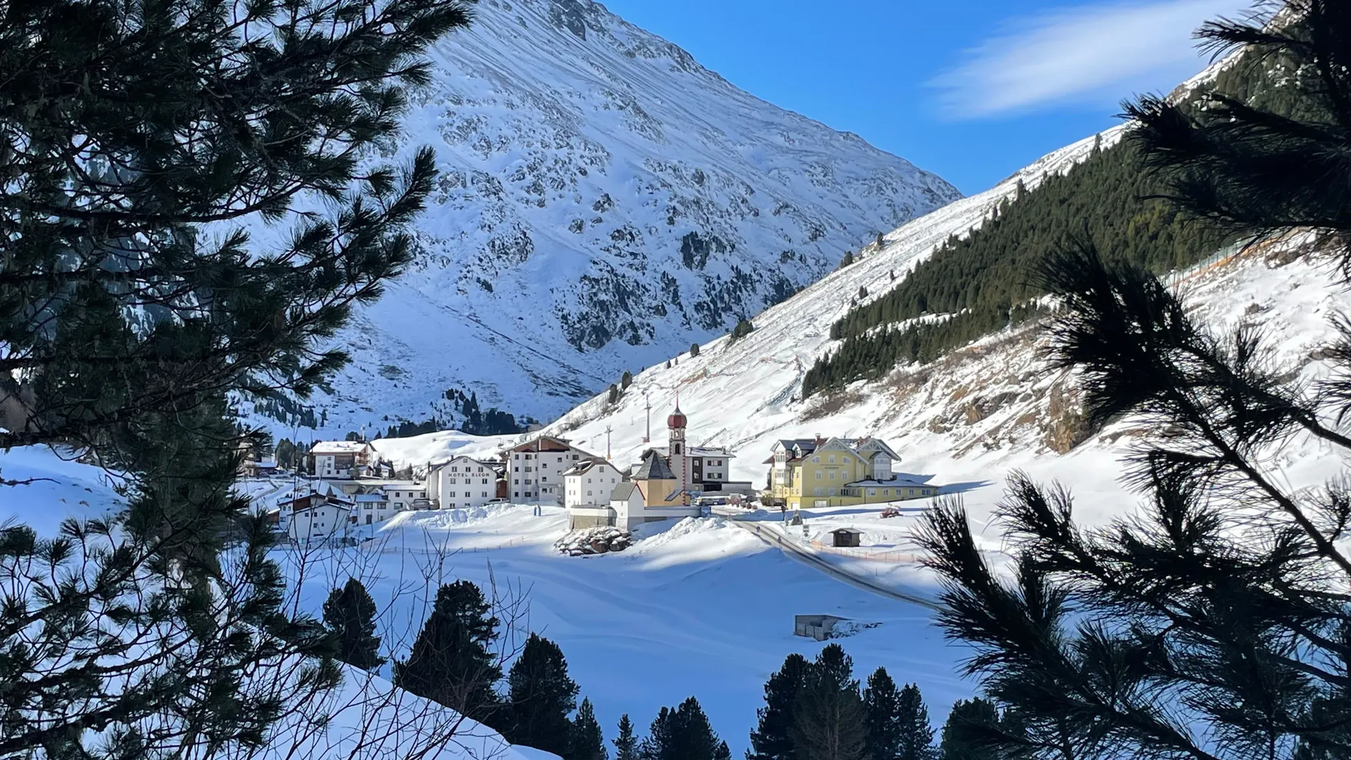Snowy mountain village with church below a snow-covered peak, framed by pine trees