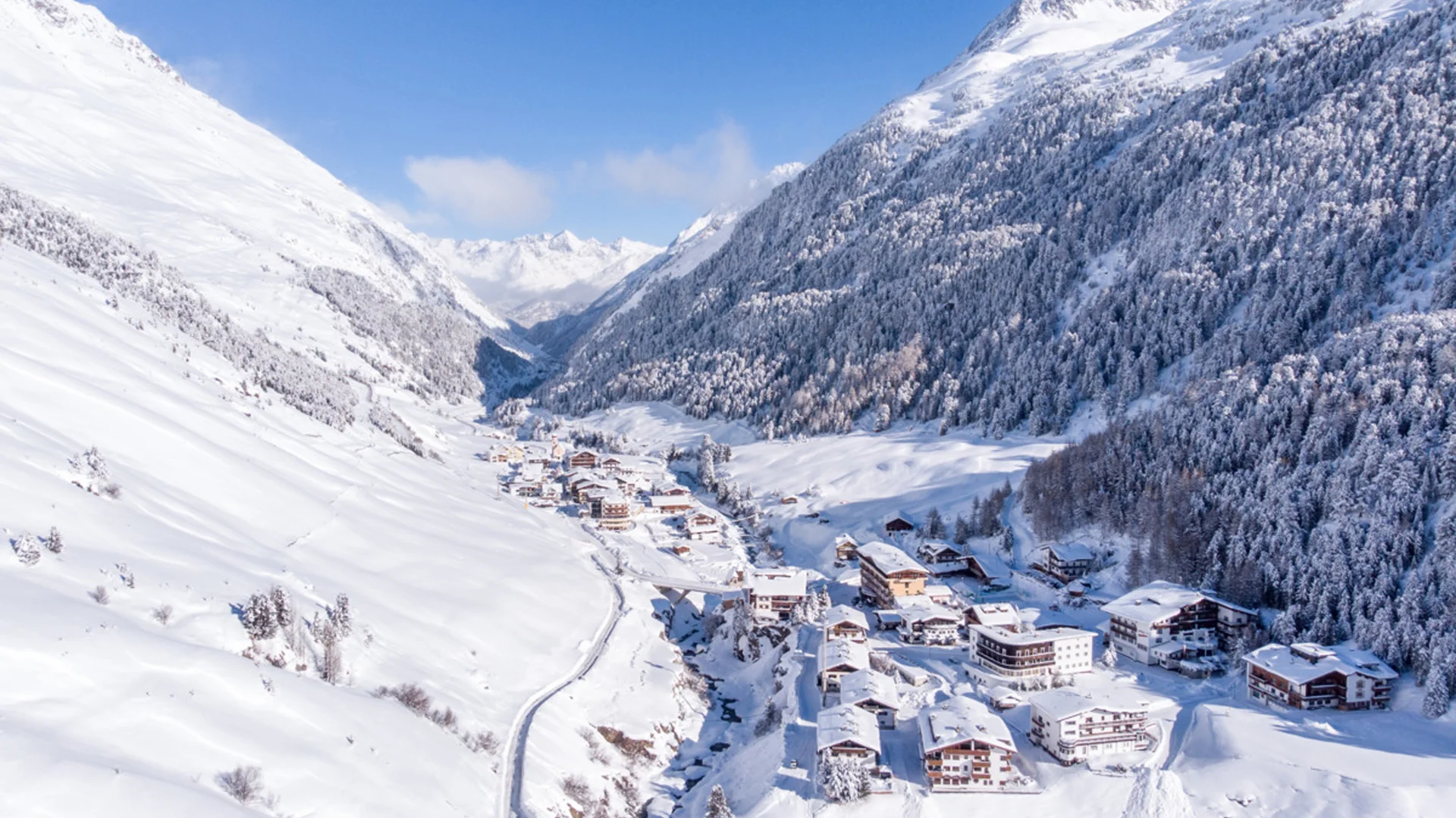 Schneebedecktes Bergdorf in einem Tal mit bewaldeten Hängen und blauem Himmel