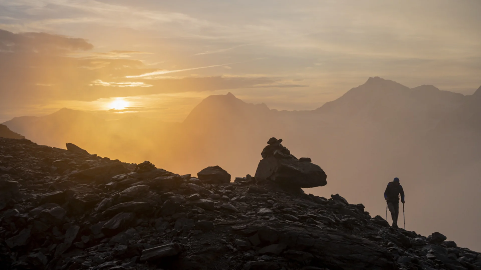 Wanderer auf felsigem Bergpfad bei Sonnenaufgang mit Nebel und Bergen