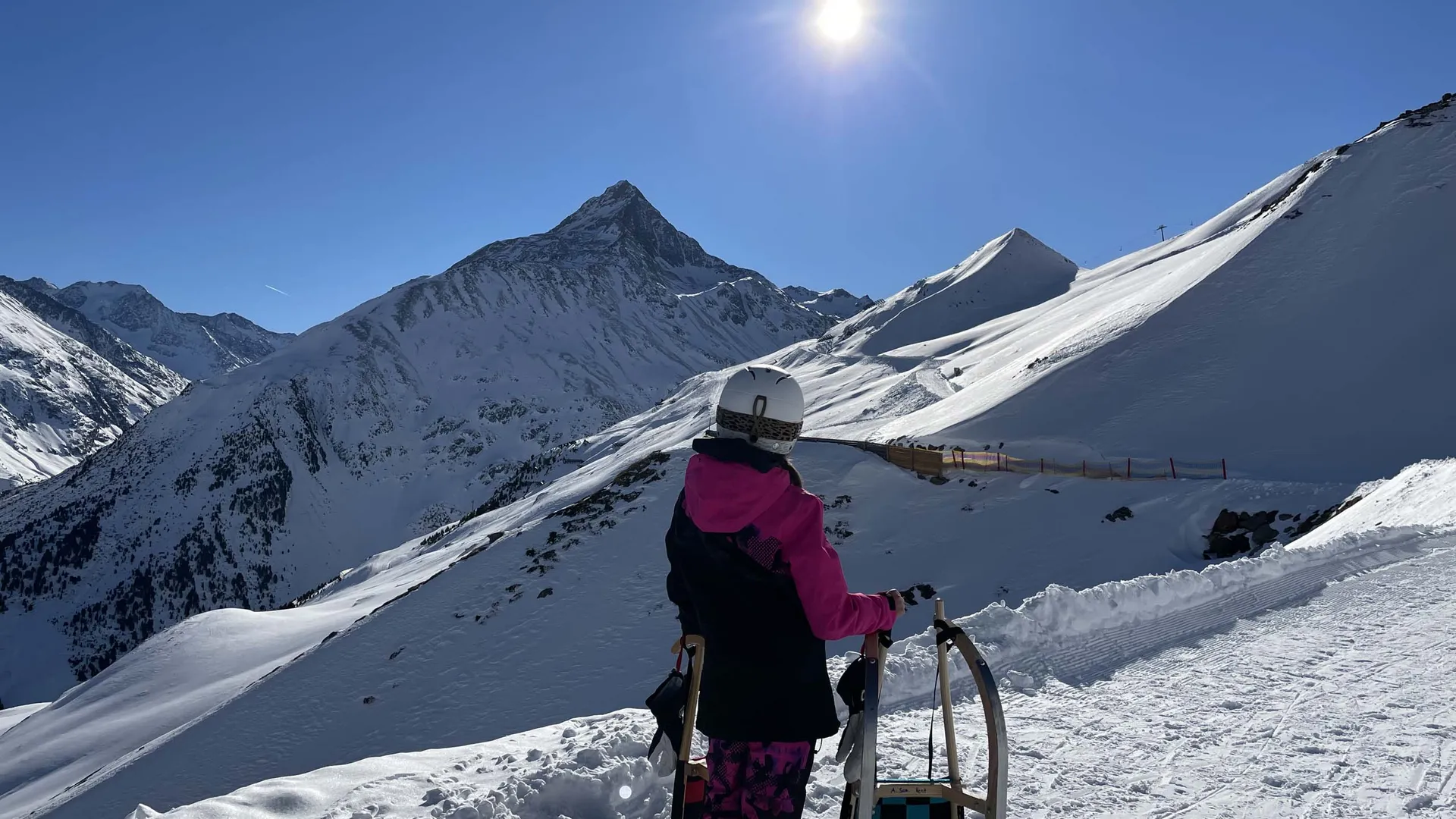 Person in pink-schwarzer Skikleidung blickt auf schneebedeckte Berge unter strahlender Sonne