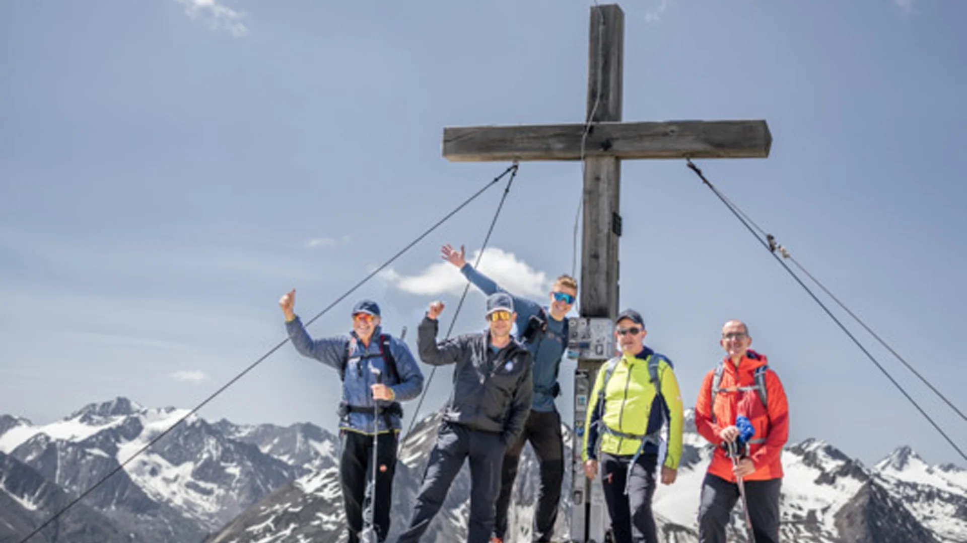 Fünf Wanderer stehen auf einem Berggipfel neben einem großen Holzkreuz