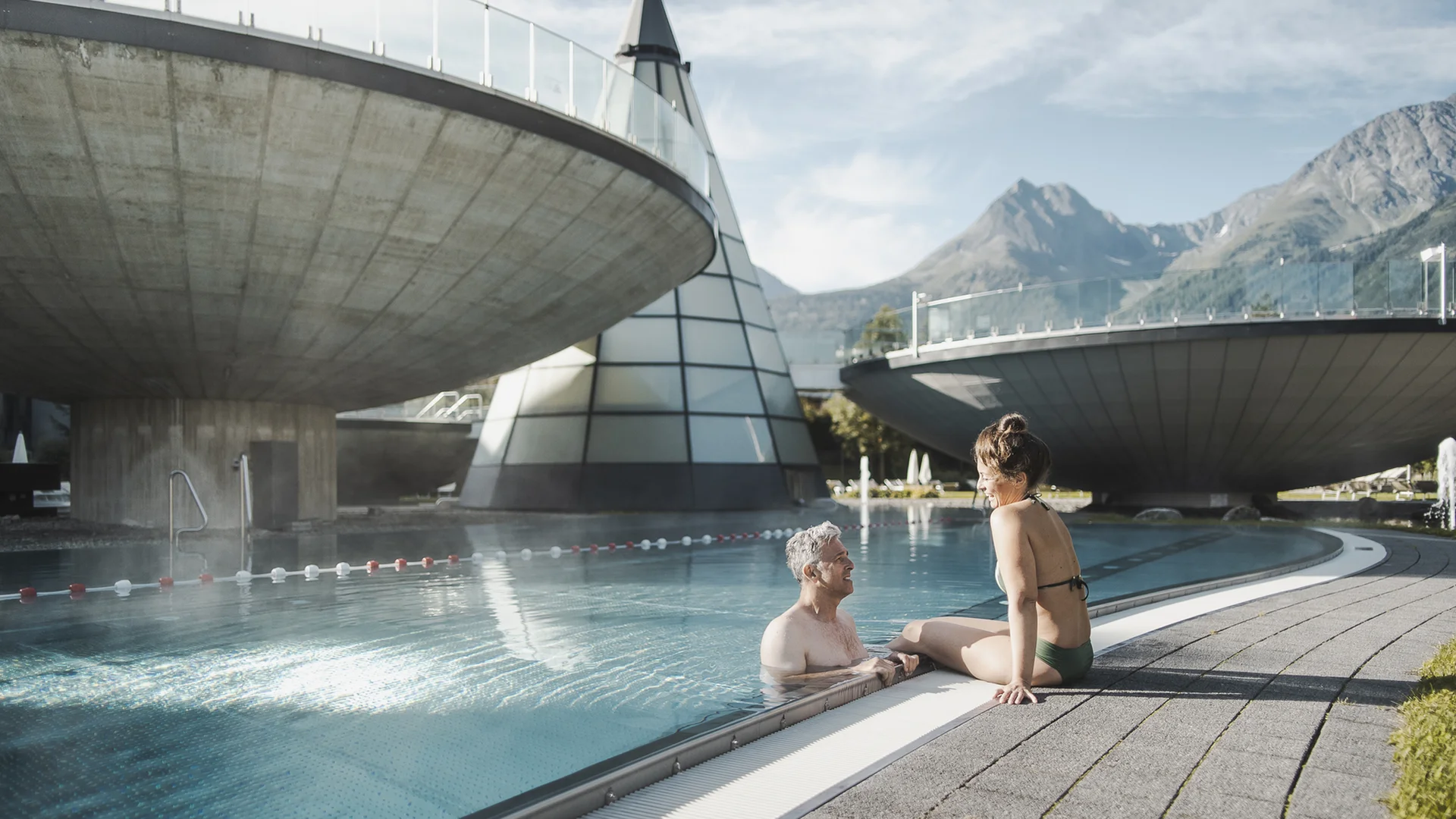 Älteres Paar entspannt am Außenpool mit moderner Architektur und Bergen im Hintergrund