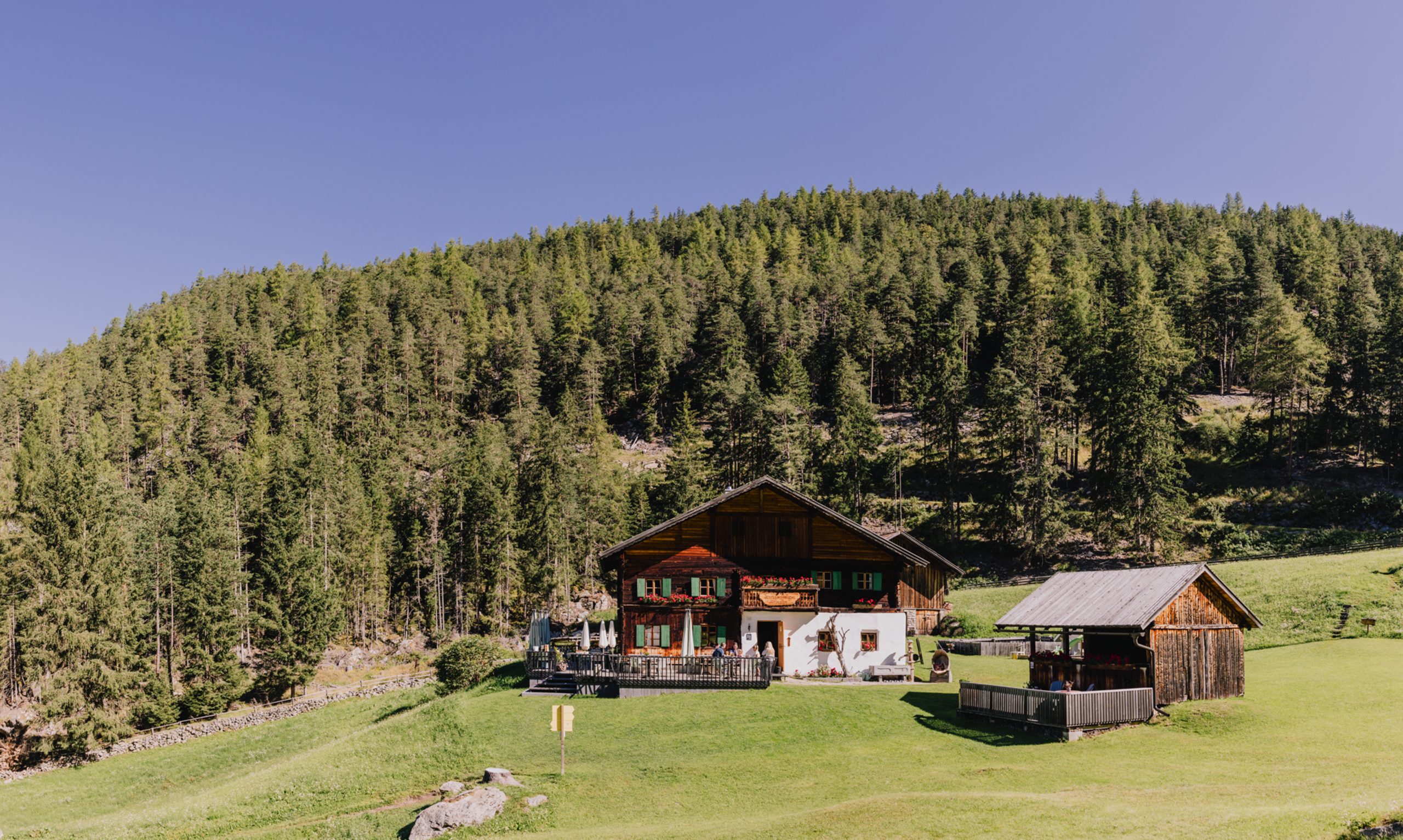 Altes Bauernhaus mit Terrasse vor bewaldetem Hügel unter klarem Himmel