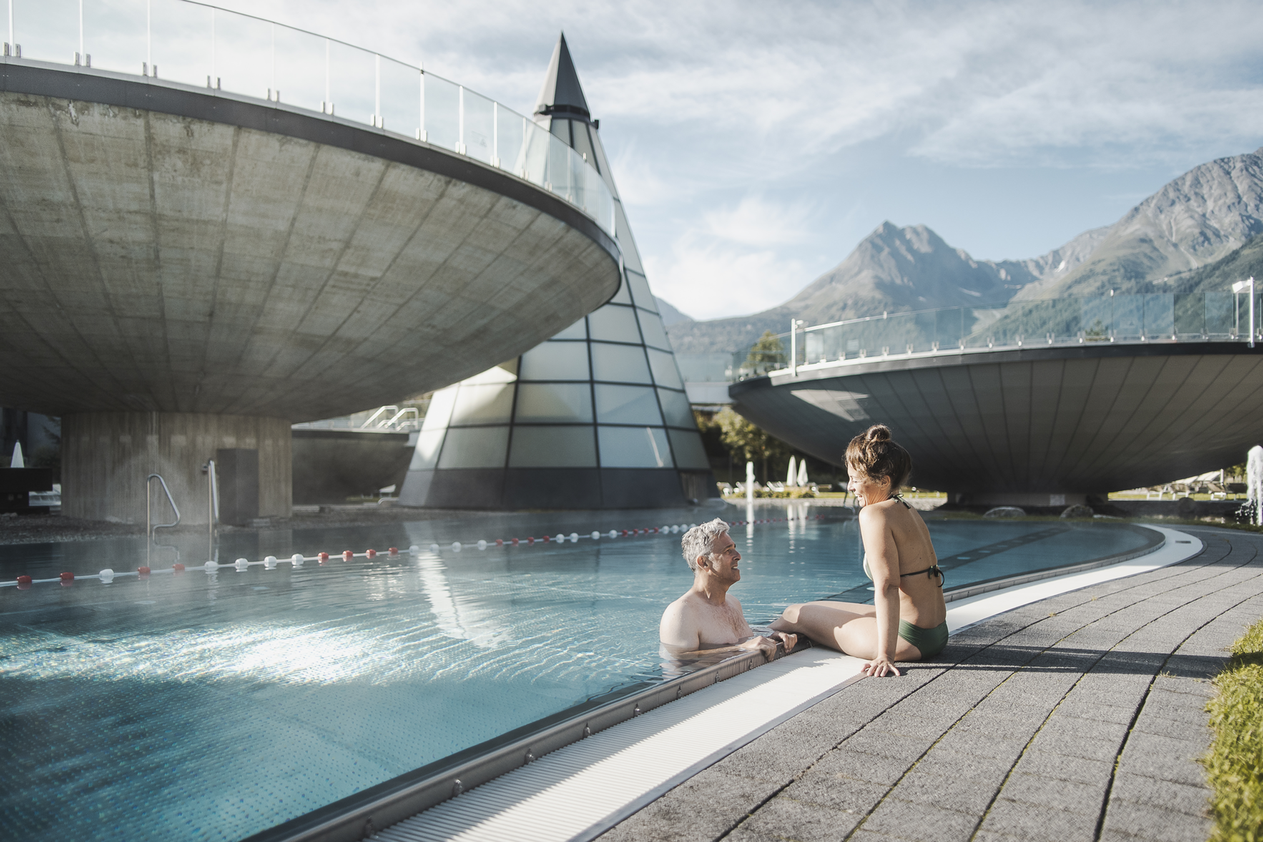 Älteres Paar entspannt am Außenpool mit moderner Architektur und Bergen im Hintergrund