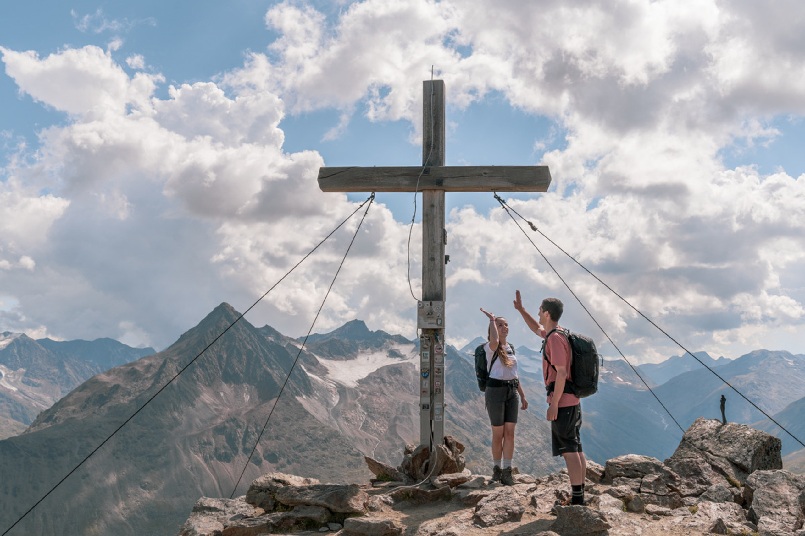 Zwei Wanderer geben sich auf Berggipfel neben großem Holzkreuz High Five