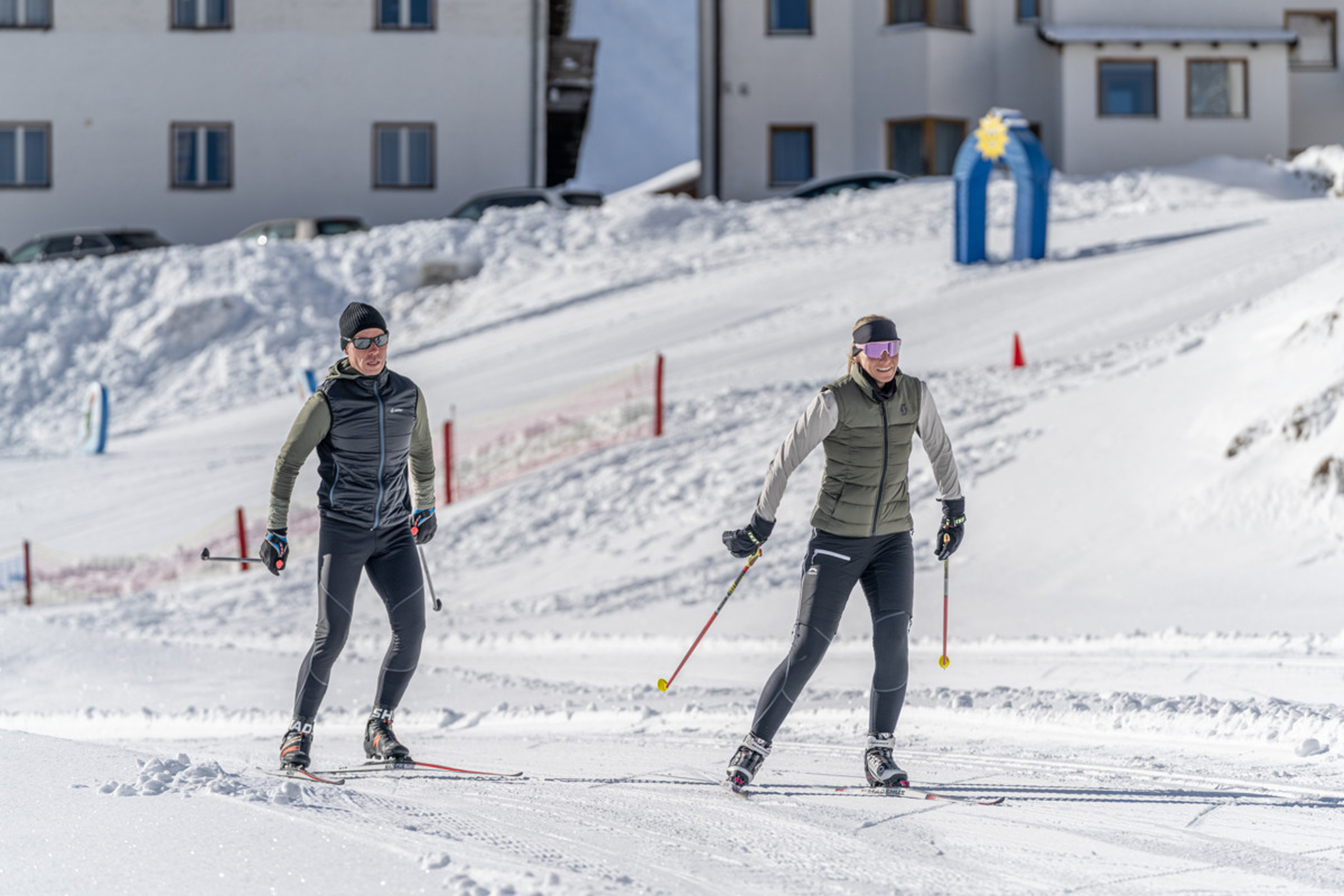 Zwei Personen beim Skilanglauf auf verschneiter Strecke vor Gebäuden