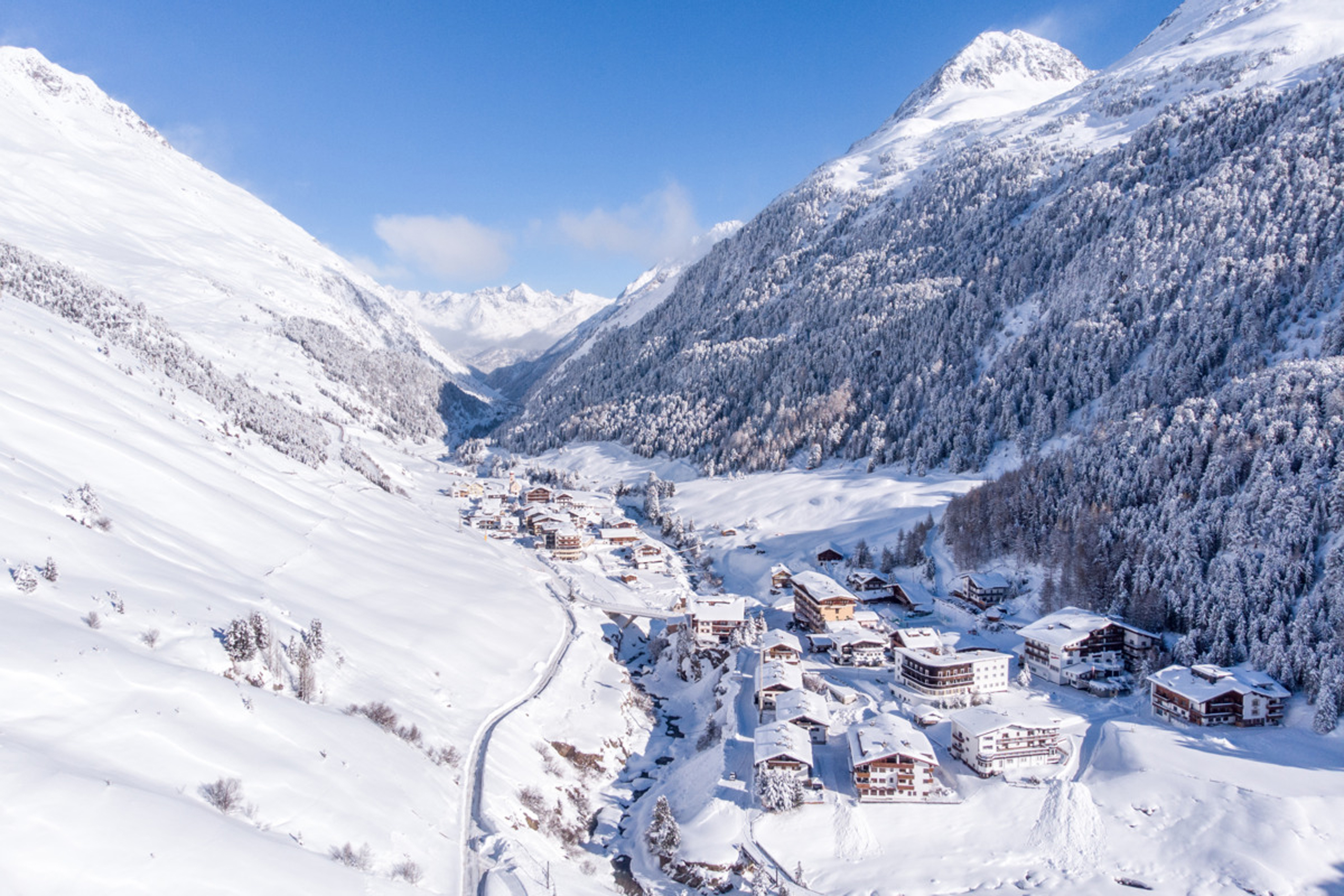 Schneebedecktes Bergdorf in einem Tal mit bewaldeten Hängen und blauem Himmel