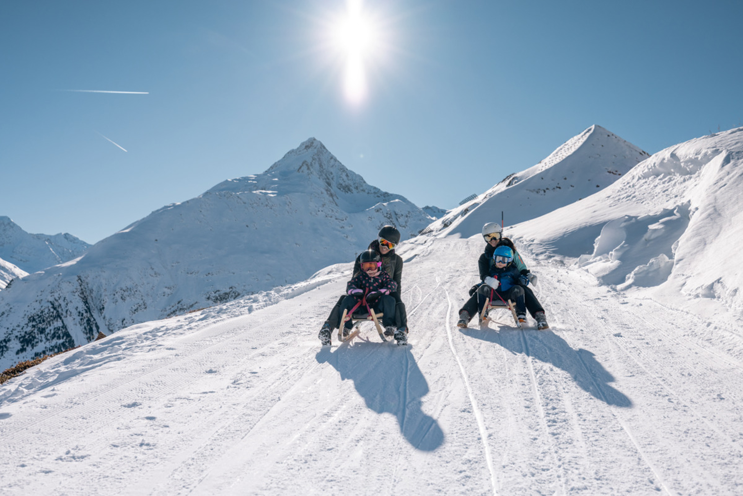 Zwei Erwachsene mit Kindern rodeln auf verschneitem Berghang bei Sonnenschein